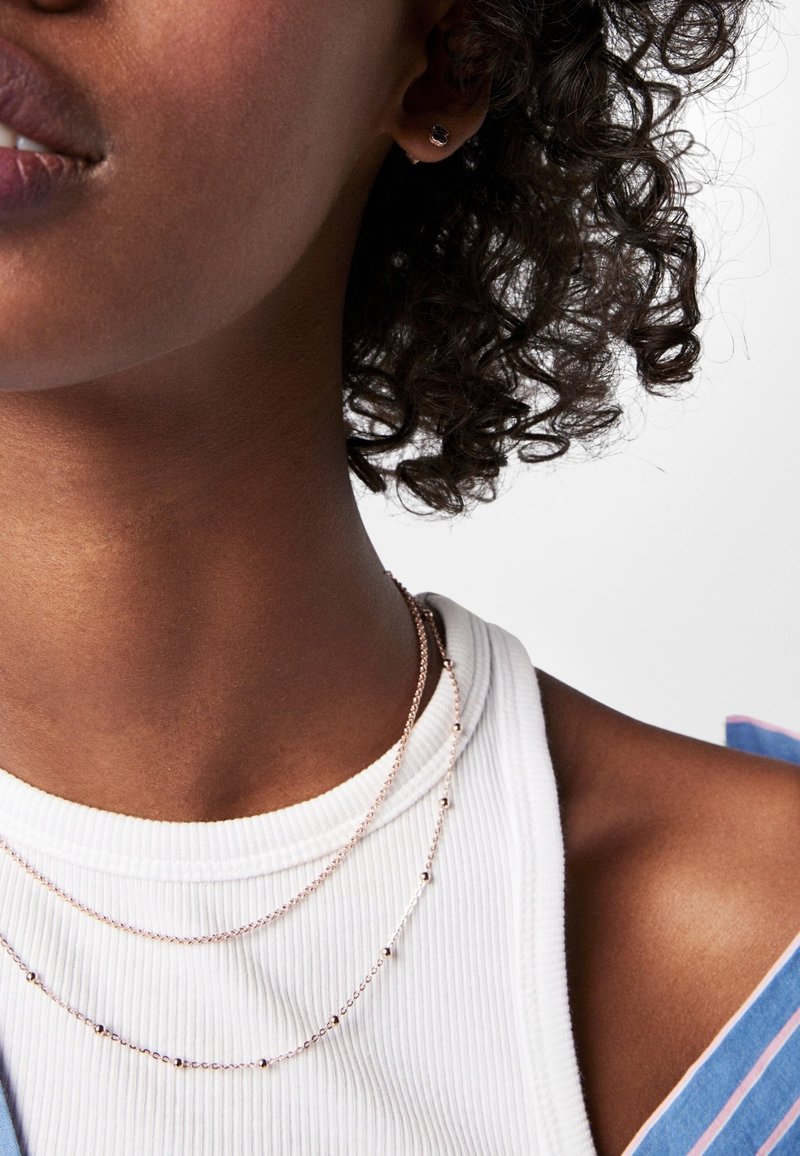 Primer plano de una persona que lleva dos delicados collares de oro y una blusa blanca de canalé debajo de una prenda a rayas azul y rosa.