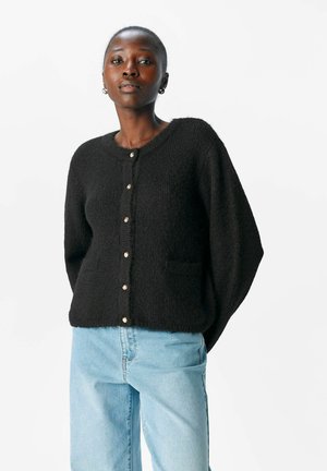 Schwarze, strukturierte Strickjacke mit rundem Hals, Knöpfen vorne und zwei Taschen. Kombiniert mit hellblauen Jeans für einen Kontrast.