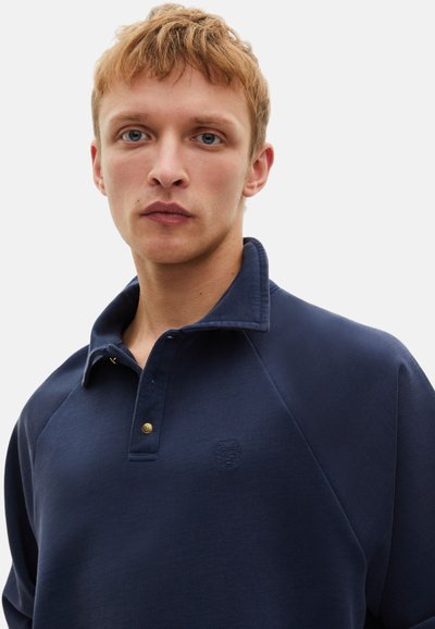 Polo shirt blu navy con patta a bottoni, colletto a coste e un ricamo logo discreto. Presenta maniche a raglan per una vestibilità rilassata.