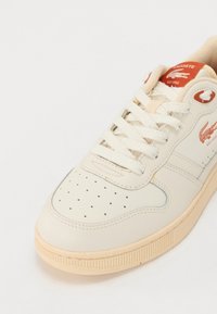 Sneaker in pelle crema con logo Lacoste rosso, punta perforata, lacci crema e suola spessa beige con dettagli testurizzati.