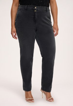Femme portant un jean noir taille haute avec une fermeture double bouton et des sandales beiges à talons hauts à brides, debout devant un fond uni.
