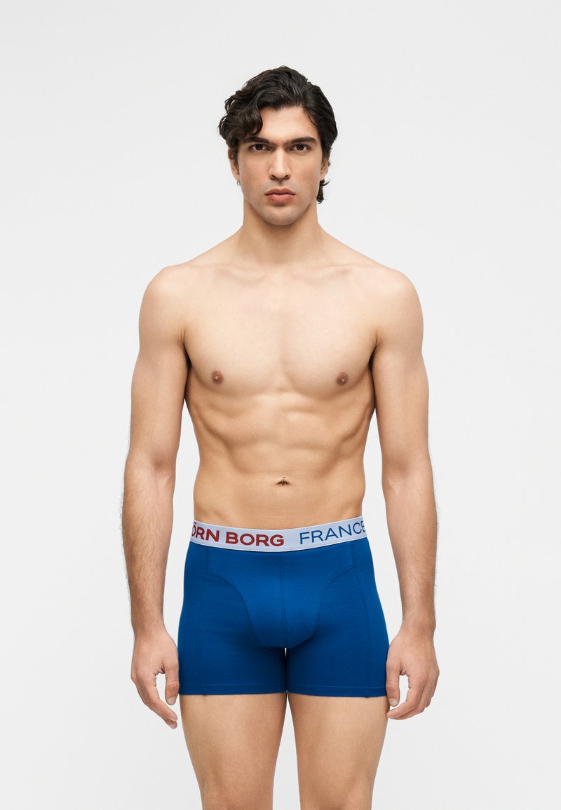 Άντρας χωρίς πουκάμισο με σκούρα μαλλιά, που φοράει μπλε μπόξερ με λευκή ζώνη που φέρει την επιγραφή «BJORN BORG FRANCE» σε λευκό φόντο.