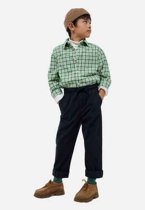 Chemise à carreaux verte et blanche, pantalon tapered bleu marine, chaussures marron à lacets, bonnet en tricot beige, col roulé blanc sous la chemise.