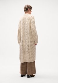 Cardigan lungo in maglia beige con motivi testurizzati e polsini a coste. Abbinato a pantaloni a quadri marroni a gamba larga e stivali col tacco marroni.