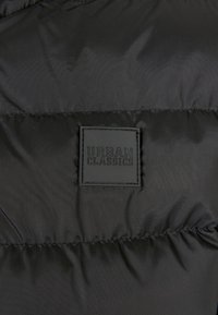 Schwarzer gepolsterter Stoff mit einem aufgenähten quadratischen Logopatch mit der Aufschrift "Urban Classics" in schwarzen erhabenen Buchstaben.