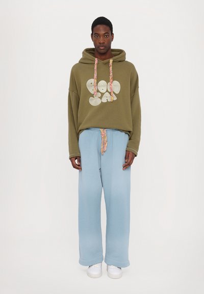 Olivenfarvet hoodie med sølv grafisk design og farverig snor. Parret med lyseblå vide sweats og hvide sneakers.