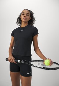 Top atlético negro con mangas cortas, que presenta un logo blanco de Nike. Combinado con pantalones cortos negros, sosteniendo una raqueta de tenis y una pelota de tenis.