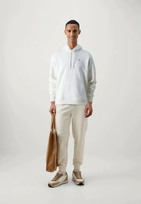 Young man in white hoodie and beige joggers holding brown tote bag, wearing beige and white sneakers, standing on plain light background.