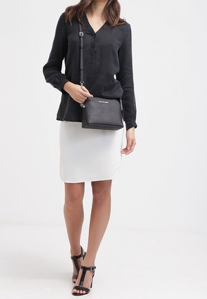 Černá crossbody taška se strukturovaným povrchem, stříbrným kováním a nastavitelným popruhem; kombinovaná s bílou sukní a černými sandály na podpatku.