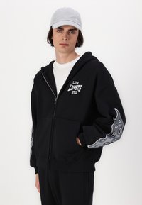 Svart zip-up hettegenser med frontlommer, med hvit tekst "LOW LIGHTS STD" og grå flamme-lignende grafikk på ermene. Myk stofftekstur.