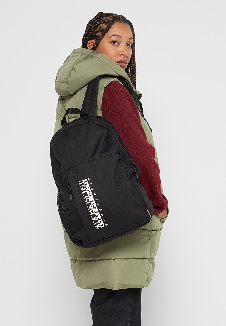 Młoda kobieta z warkoczami, ubrana w zieloną pikowaną kamizelkę i bordowy sweter, nosi czarny plecak Napapijri na jednym ramieniu.