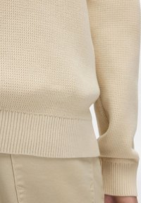 Beige gestrickter Pullover mit strukturiertem Stoff und gerippten Bündchen, kombiniert mit hellbeigen Hosen, die ein dezentes, monochromatisches Farbschema zeigen.