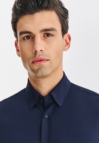 Granatowa koszula zapinana na guziki z ostrym kołnierzykiem i gładką fakturą. Posiada guziki z przodu oraz dopasowany krój. Bez wzorów i zdobień.