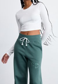 Bílý krátký top s dlouhým rukávem a malým logem na hrudi, doplněný zelenými tepláky se stahovací šňůrkou a vyšitým názvem značky.