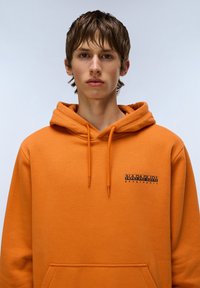 Felpa arancione realizzata in tessuto morbido. Presenta una tasca a marsupio e un piccolo logo stampato sul lato sinistro del petto. Il cappuccio è dotato di lacci.