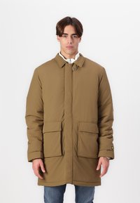 Bruine gewatteerde jas met een hoge kraag, twee voorzakken en een klein logo op de mouw. Soepele textuur, iets oversized pasvorm.