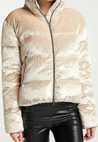 Chaqueta acolchada color crema con una tela suave y brillante, cierre de cremallera y capucha. Presenta un acolchado horizontal y puños extragrandes.