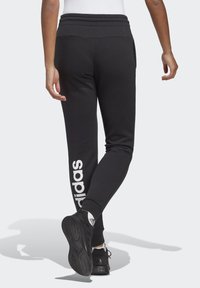 Fekete joggingnadrág rugalmas derékpánttal, oldalsó zsebekkel és fehér "adidas" logóval a bal lábon. Puha anyag és szűkebb szabás.