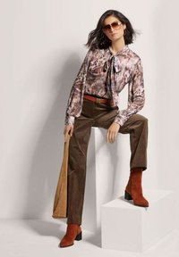 Bruine blouse met patroon en strik, gecombineerd met bruine corduroy broek en roestkleurige suède enkelboots. Accessoires zijn onder andere een zonnebril.