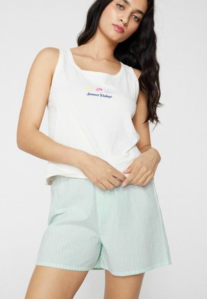 Kobieta w białej bluzce bez rękawów z napisem „Summer Feelings” i ikonami, w połączeniu z jasnoszortami w zielone paski na białym tle.