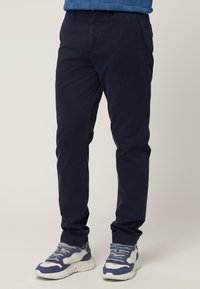 Chino blu navy realizzati in tessuto misto cotone, progettati con una vestibilità slim, dotati di tasche laterali e una texture morbida, abbinati a sneaker grigie e blu.