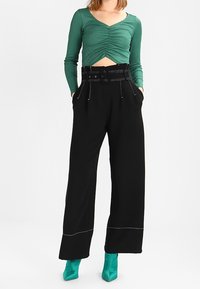Green long-sleeve crop top with ruched detailing, paired with high-waisted black wide-leg trousers featuring white stitching and a belted waist.