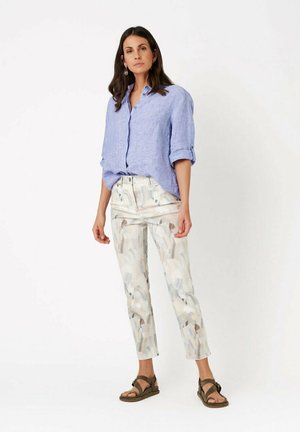 Woman wearing light blue rolled-up sleeve shirt, abstract patterned beige pants, and brown sandals standing against white background.