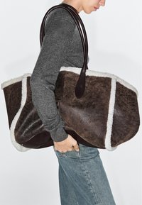 Person in gray sweater and jeans carrying large brown shearling tote bag with dark handles on shoulder.
