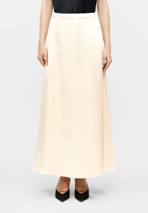 Γυναίκα που φοράει μακριά, κρεμ φούστα με ελαστική μέση, συνδυασμένη με μαύρα παπούτσια με μυτερή μύτη και μαύρο πάνω μέρος.