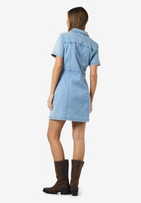 Vestido de ganga curto em azul claro com mangas curtas, colarinho e cintura justa. Usado com botas castanhas até ao joelho.