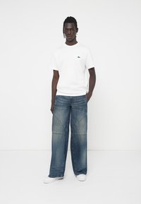 Camiseta blanca de algodón con un pequeño logo aguamarina, combinada con unos vaqueros de mezclilla azul de pierna ancha que presentan un detallado distintivo en las costuras. Zapatillas blancas completan el look.