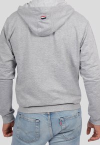 Sudadera gris con capucha, hecha de material suave, que presenta una pequeña etiqueta bordada en rojo, blanco y azul en la parte trasera, combinada con jeans azules.