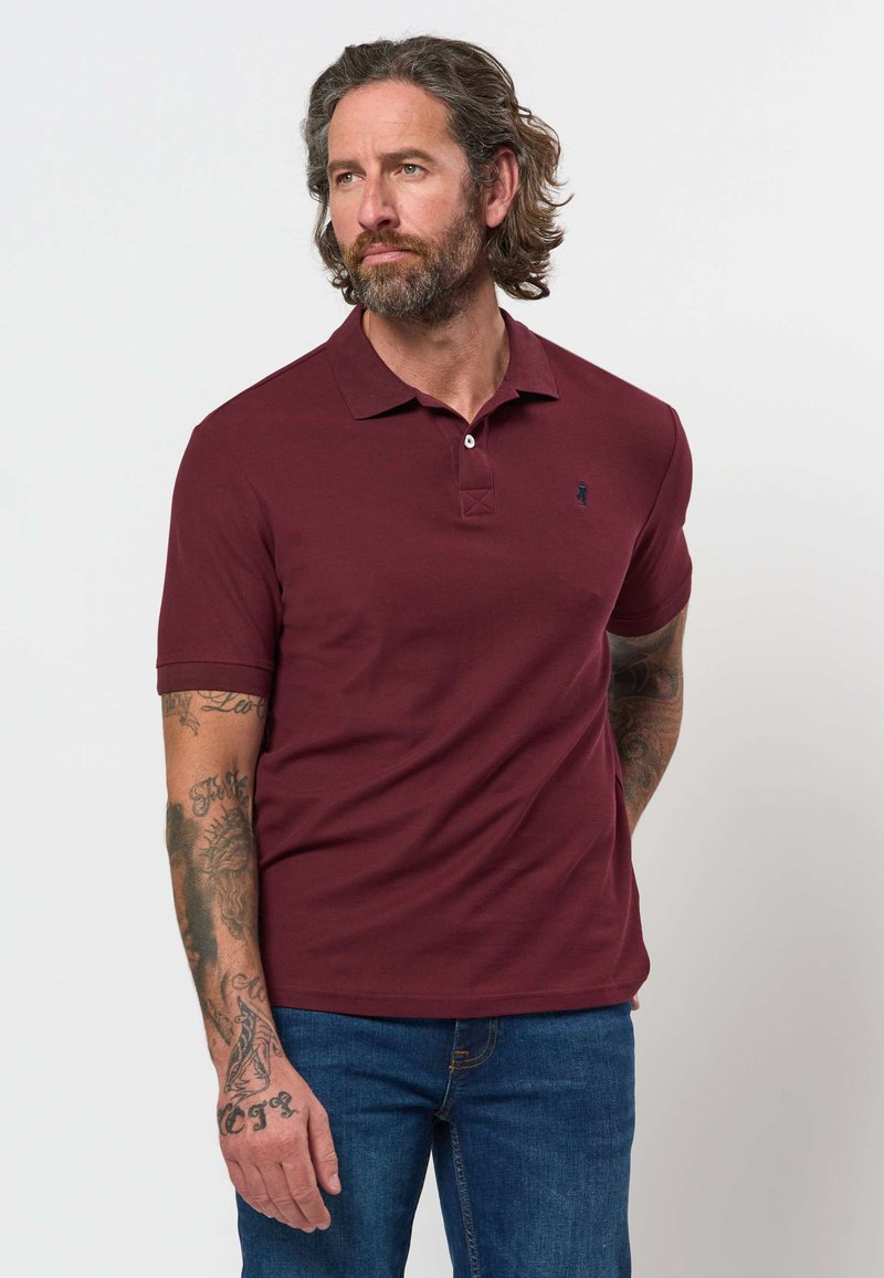 Homme avec une barbe et des tatouages portant un polo grenat et un jean bleu, debout devant un fond clair uni.