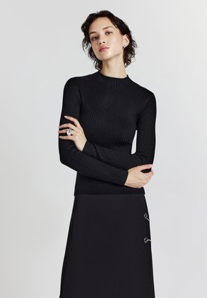Femme aux cheveux courts et foncés portant un haut noir à manches longues en côtes et une jupe noire avec deux détails en anneaux argentés, les bras croisés sur la poitrine.