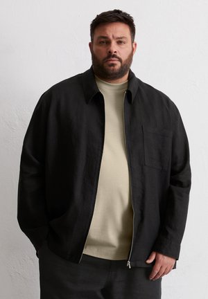 Black linen jacket with a collared design, front zipper, and single chest pocket, layered over a beige short-sleeve shirt.