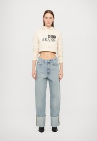 Zkrátená béžová mikina s čiernym potlačou, spárovaná s svetlo modrými širokými džínsami s rozstrapkanými okrajmi a žltými švami.