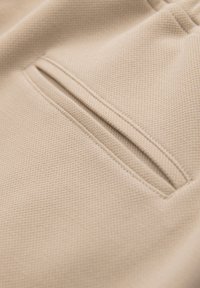 Gros plan sur un tissu beige texturé avec une poche fendue horizontale double surpiqûre, montrant une couture détaillée et un bord de ceinture élastique.