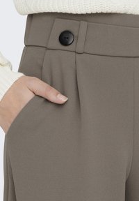 Beige-texturoitu housut, joissa on kapeneva muoto, mustalla napilla suljettuna ja sivutaskuilla, yhdistettynä kermaiseen neuleeseen.