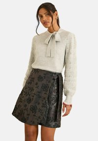 Világosszürke kötött pulóver masnival a nyakkivágásnál és bonyolult mintázatú ujjakkal, fekete virágmintás szoknyával párosítva.