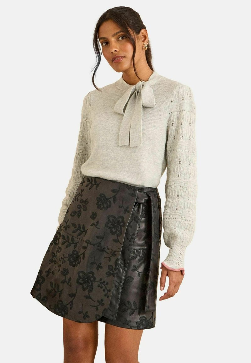 Világosszürke kötött pulóver masnival a nyakkivágásnál és bonyolult mintázatú ujjakkal, fekete virágmintás szoknyával párosítva.