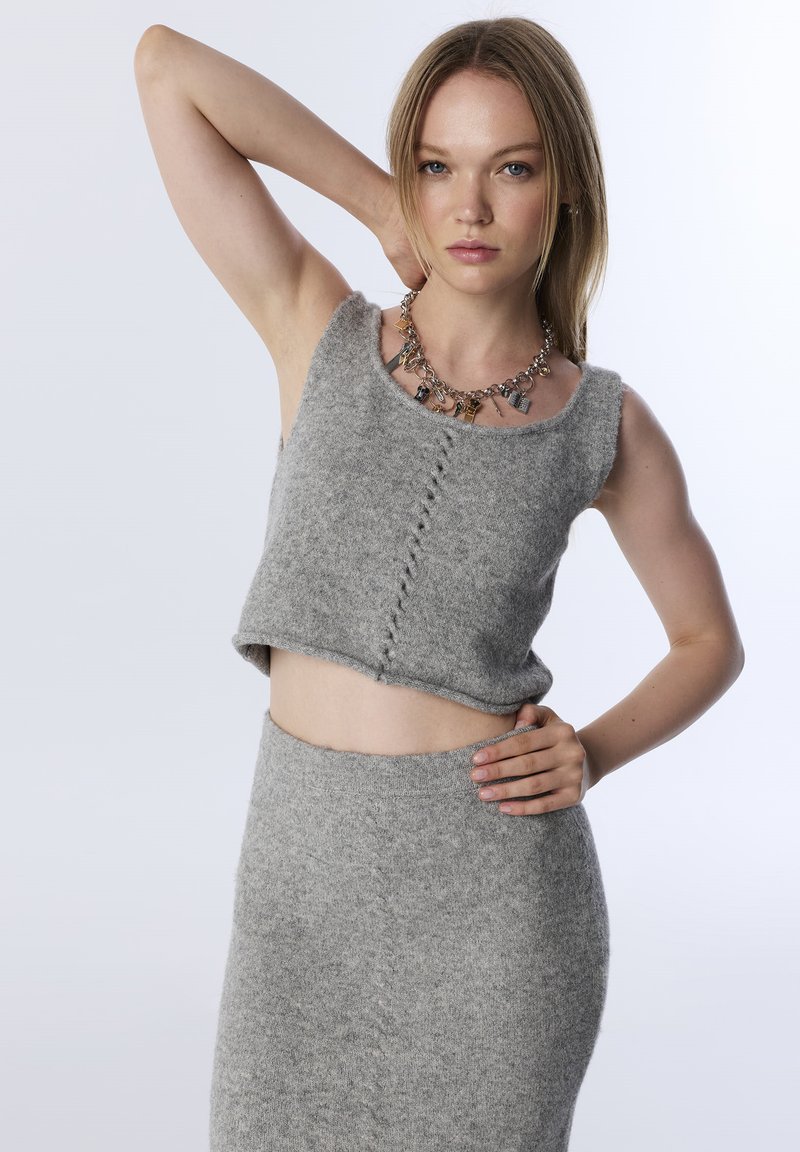 Młoda kobieta w szarym dzianinowym topie na krótki rękaw i spódnicy, nosząca naszyjnik z charmsami, z jedną ręką na biodrze, a drugą za głową.