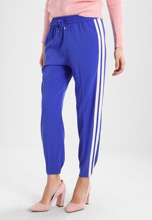 Ben iført blå træningsbukser med hvide sideliner, kombineret med lyserøde høje hæle og en lyserød sweater synlig ved taljen.