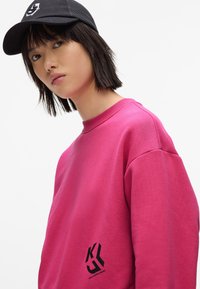 Felpa fucsia con colletto rotondo e spalle ribassate, caratterizzata da una stampa nera del logo sul lato sinistro e da un tessuto testurizzato. Un cappellino in rete nera completa il look.