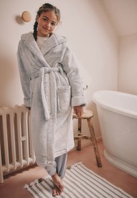 Accappatoio grigio chiaro, soffice, con cappuccio, cintura annodata e tasche laterali; indossato sopra pantaloni del pigiama a righe in un ambiente bagno.