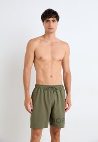 Olivengrønne badeshorts med elastisk talje og snøring. Har et dristigt sort tekstdesign på venstre ben. Glat tekstur.