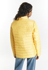 Gul vadderad jacka med horisontell sömnad, hög krage och något figursydd form. Bärs över denimjeans, vilket visar en lätt, strukturerad yta.