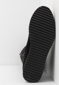 Semelle de botte noire avec des rainures diagonales, positionnée verticalement contre un mur d'angle blanc et le sol.