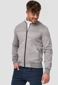 Siva laža suđena bomber jakna s crnim patentnim zatvaračem, rebrastim manžetama i rubom. Ima minimalistički dizajn s teksturiranom površinom.