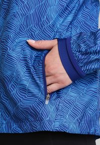 Veste de sport bleue avec un motif de vagues, dotée d'une poche zippée et d'accents en maille aux poignets. Texture légère et lisse.