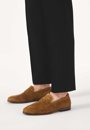 Rudi zomšiniai mokasinai dėvimi su juodomis trumpintomis kelnėmis, parodyti iš šono prieš paprastą šviesią foną.